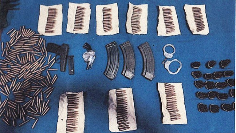 Confiscated weapons and ammunition including T-56 rounds, magazines, a micro firearm, a revolver, handcuffs, and army uniforms displayed on a table