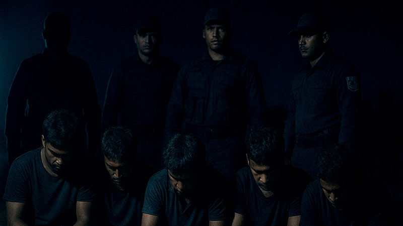 Five arrested men (suspects) sitting with their heads bowed under dim light with security officers standing behind them
