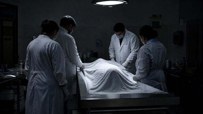 Four Judicial Medical Officers inspect a covered body in a dark examination room during a post-mortem investigation